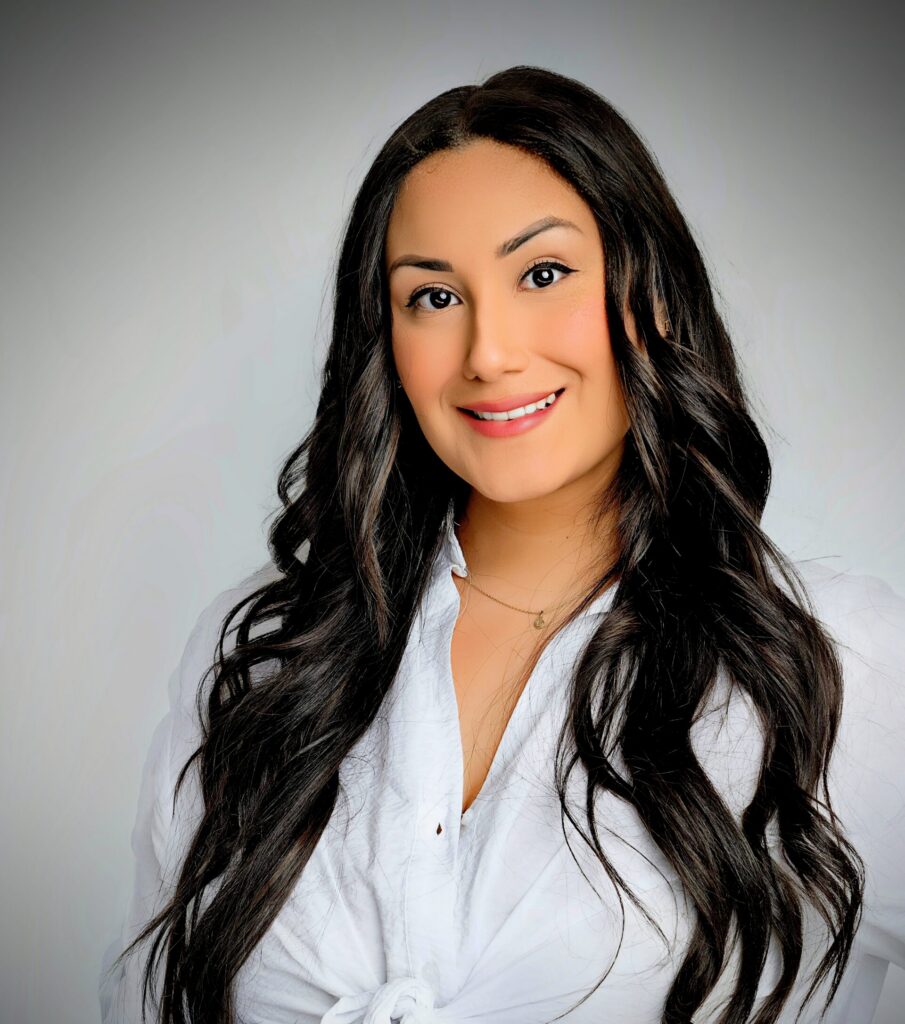 Woman with long wavy dark hair, wearing a white blouse, smiling in front of a plain light background.