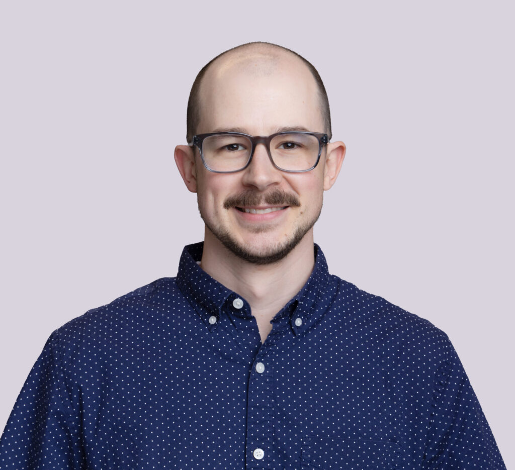 A man with glasses, a mustache, and a beard wearing a navy blue short-sleeve button-up shirt with small white dots, standing against a light gray background.