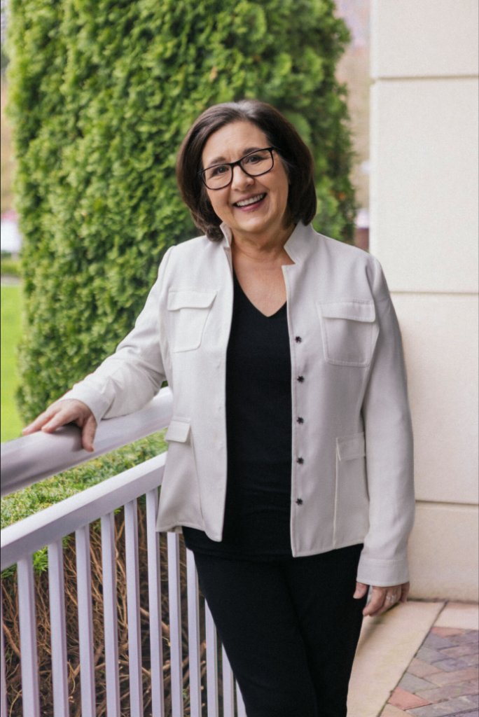 A woman with dark hair and glasses stands outdoors, smiling, wearing a white jacket over a black shirt and pants, with greenery and a building in the background.