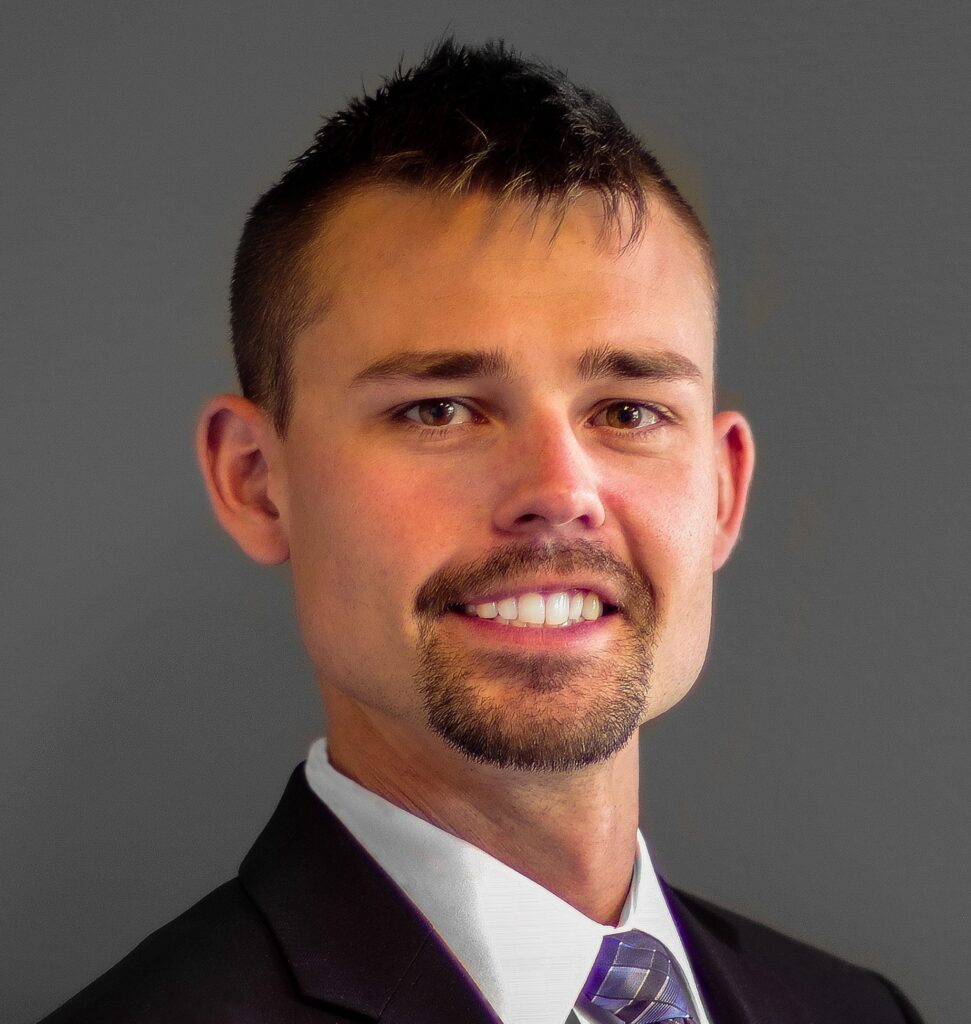 A man with short dark hair, a goatee, and a mustache smiles at the camera, wearing a suit and striped tie, against a gray background.