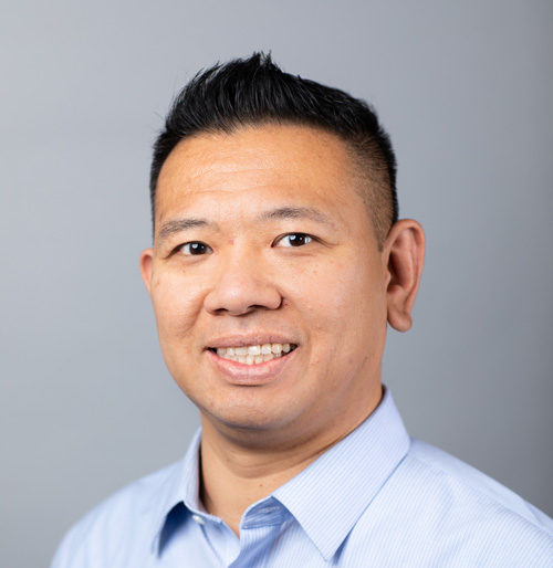A man with short black hair wearing a light blue collared shirt, smiling at the camera against a plain gray background.