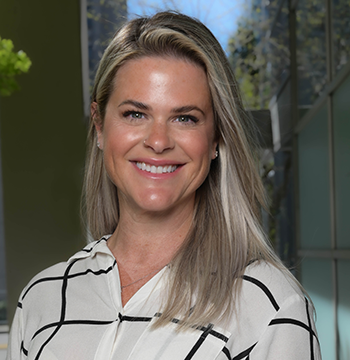 A woman with long blonde hair, wearing a white blouse with black stripes, smiles at the camera in an indoor setting with natural light and greenery in the background.
