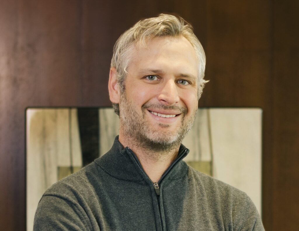 A man with gray hair and beard, wearing a dark zip-up sweater, stands smiling in front of a wooden wall and abstract artwork.