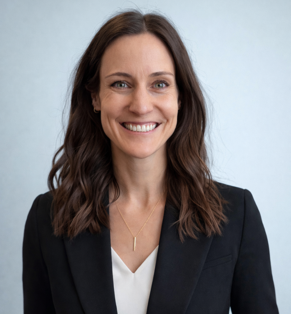 A woman with wavy brown hair, wearing a black blazer over a white top and a gold necklace, smiles at the camera against a plain light background.
