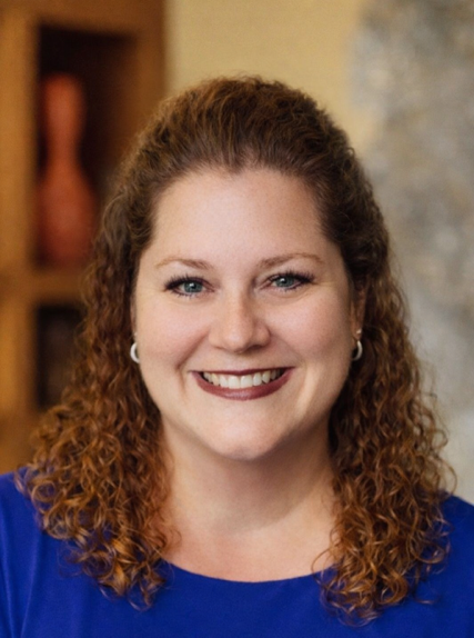 A woman with curly auburn hair, wearing a blue top and silver hoop earrings, smiles at the camera; blurred indoor background.
