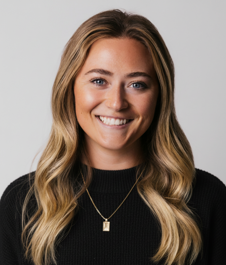 A woman with long blonde hair, wearing a black top and a gold necklace, smiles at the camera against a plain light background.