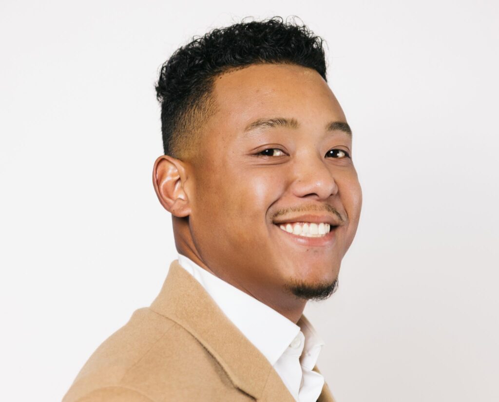 A man with short curly hair and a trimmed goatee smiles at the camera, wearing a tan blazer over a white collared shirt, against a plain white background.
