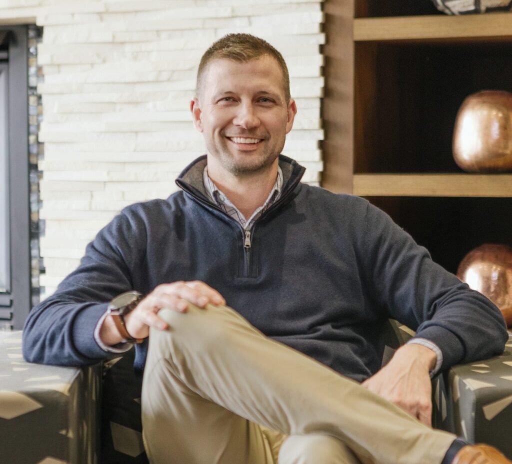 A man in a navy sweater and khaki pants sits on an armchair, smiling, with one leg crossed over the other. A light stone wall and shelves are in the background.