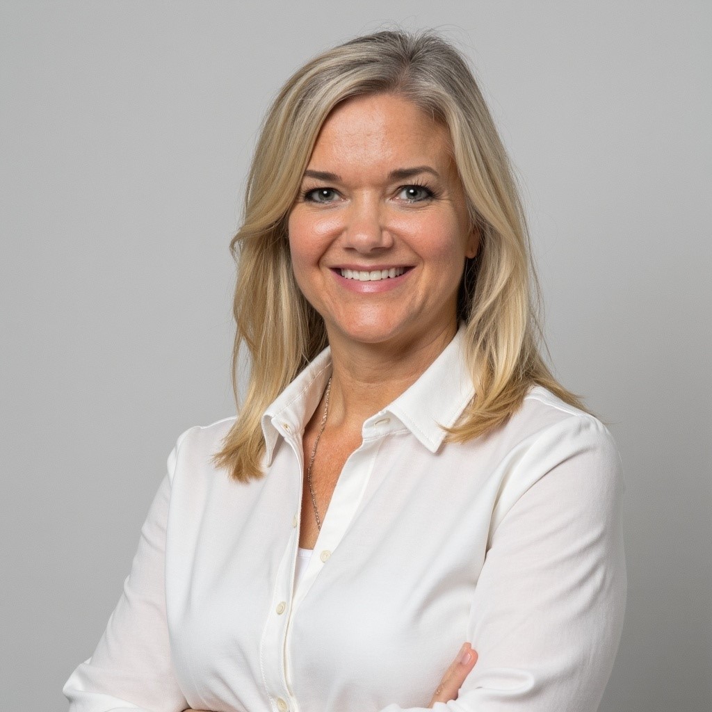 A woman with blonde hair wearing a white button-up shirt stands smiling with her arms crossed against a plain light gray background.
