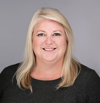 A woman with long blonde hair wearing a dark top is smiling at the camera against a plain gray background.