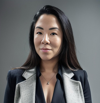 A woman with long, dark hair wearing a black blouse and a gray blazer stands in front of a plain gray background.