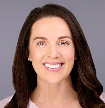 Woman with long brown hair and light skin smiling at the camera, wearing a light pink top, against a plain gray background.