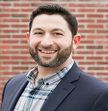 A man with short dark hair and a beard, wearing a plaid shirt and dark blazer, stands in front of a brick wall and smiles at the camera.