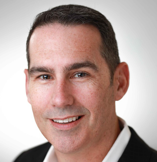 A man with short dark hair, light skin, and wearing a dark suit jacket over a white shirt, smiling at the camera against a plain light background.