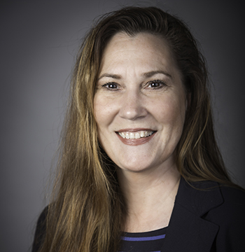 A woman with long brown hair smiles at the camera, wearing a dark blazer and striped top, against a plain dark background.