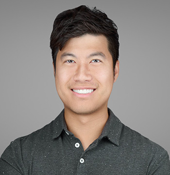 Smiling man with dark hair wearing a dark button-up shirt posed in front of a plain gray background.