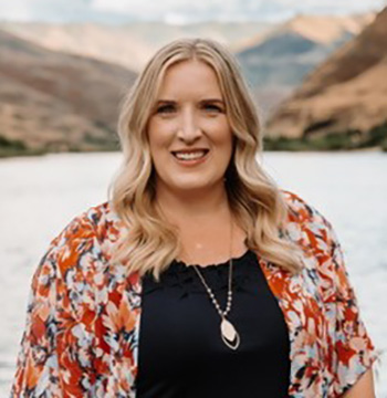 A woman with long blonde hair, wearing a black top and floral shawl, stands outdoors by a river with hills in the background.