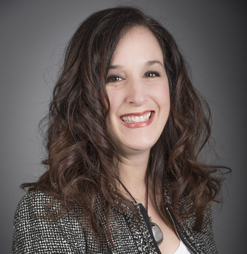 Smiling woman with long, wavy brown hair wearing a black and white blazer and a necklace, posed against a plain gray background.