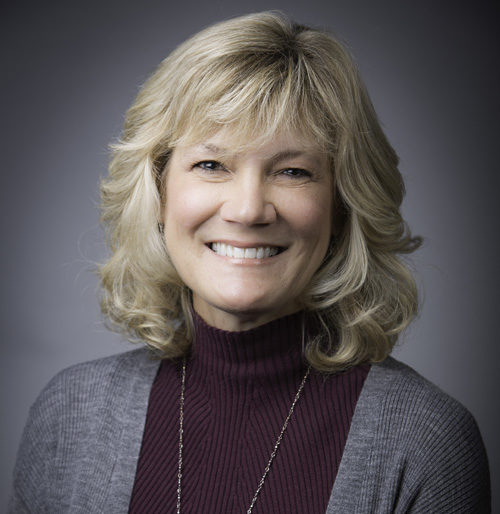 A woman with shoulder-length blonde hair smiles at the camera, wearing a maroon turtleneck and a gray cardigan in front of a plain background.