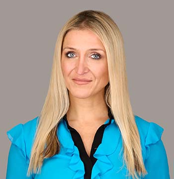 Woman with long blonde hair wearing a bright blue blouse over a black top, standing against a plain gray background.