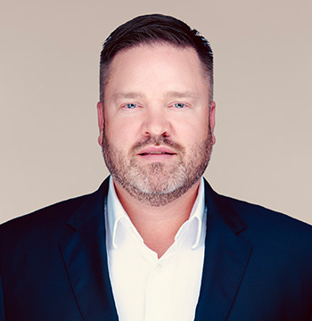 A man with short brown hair and a beard, wearing a dark suit jacket and white collared shirt, poses against a plain light background.