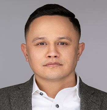Man with short dark hair wearing a grey blazer and white dress shirt, facing forward with a neutral expression against a plain background.