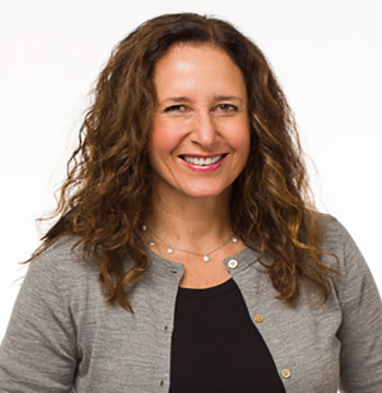 A woman with long curly brown hair, wearing a gray cardigan over a black top, smiles at the camera against a plain white background.