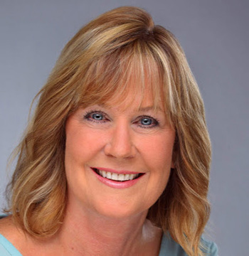 Smiling woman with shoulder-length blonde hair and blue eyes, wearing a light blue top, photographed against a plain gray background.