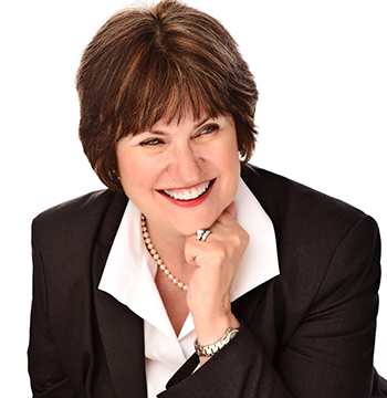 Woman wearing a dark blazer, white blouse, and pearl necklace, smiling with hand resting on chin against a white background.