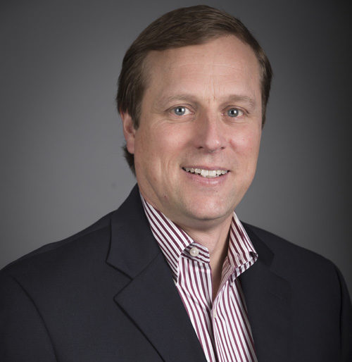 A man with short light brown hair, wearing a dark suit jacket over a red and white striped shirt, poses against a plain dark gray background.