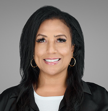 Woman with long dark hair, wearing hoop earrings, a black blouse over a white top, smiling at the camera against a plain gray background.