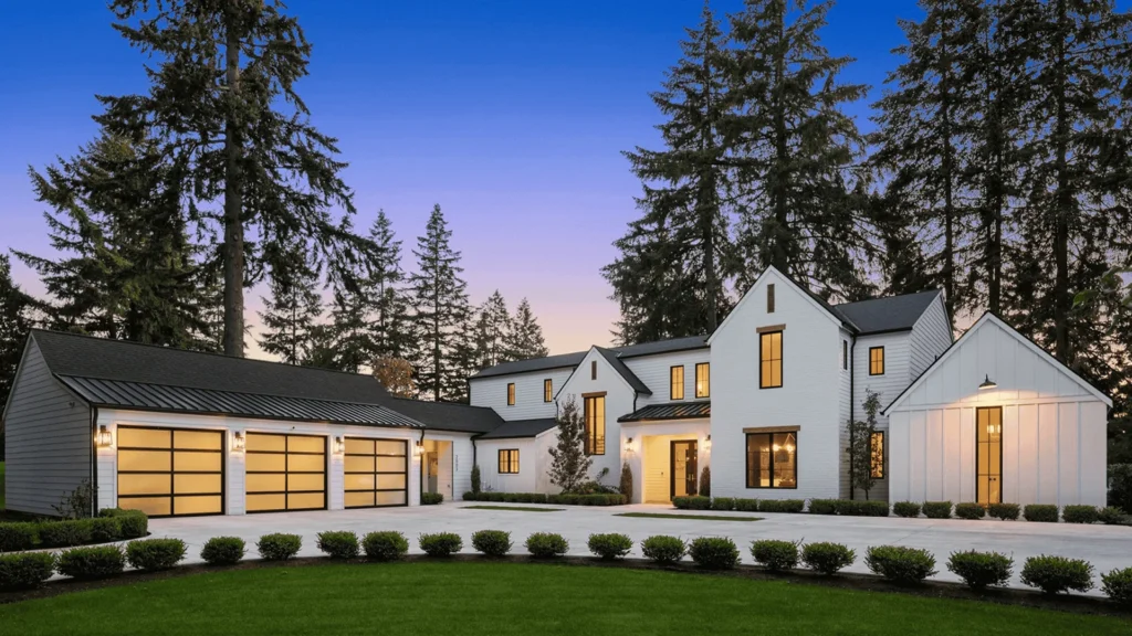 Modern two-story house with white exterior, large windows, and a three-car garage, surrounded by neatly trimmed bushes and tall trees at dusk.