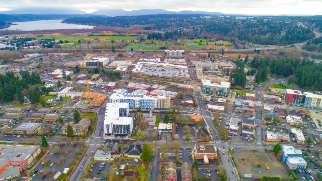 Aerial view of a suburban city with residential and commercial buildings, roads, parking lots, trees, and a river in the background.