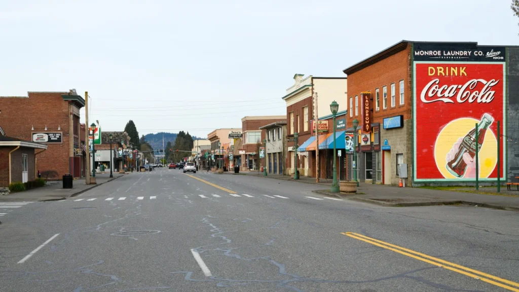 A quiet small-town street with historic buildings and a large Coca-Cola mural on one wall, under an overcast sky.