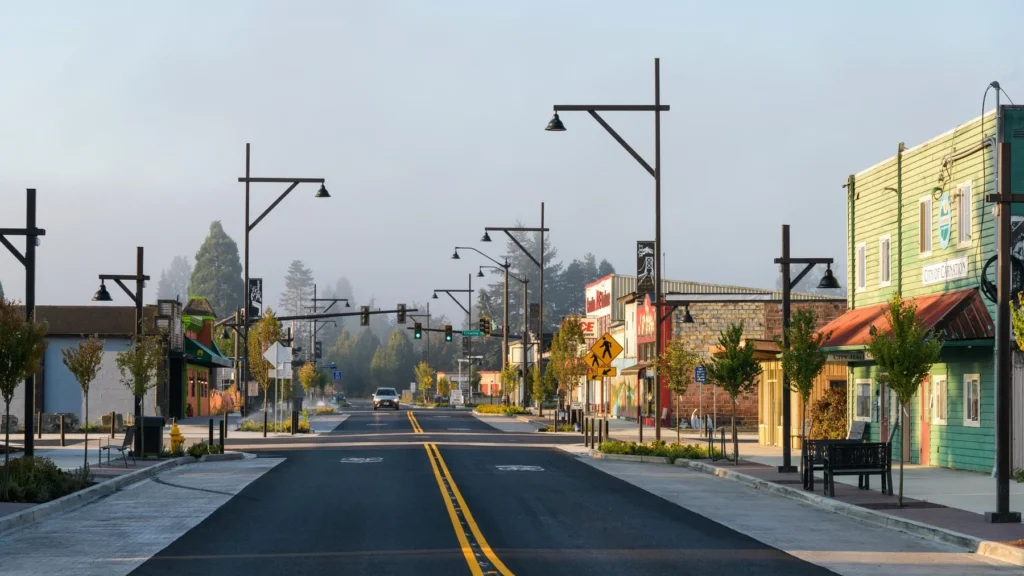 A wide, empty street in a small town with storefronts and streetlights on both sides, trees lining the sidewalks, and a hazy sky in the background.