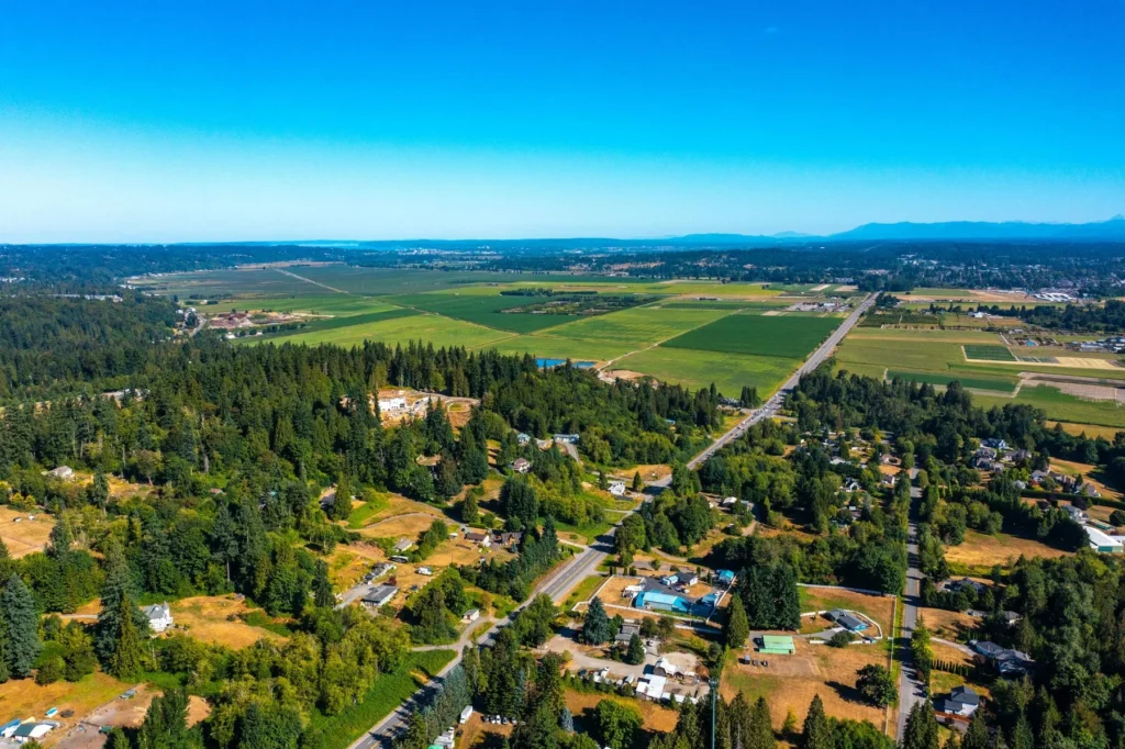 Aerial view of a rural area with forests, scattered houses, open fields, and a straight road running through the landscape under a clear blue sky.