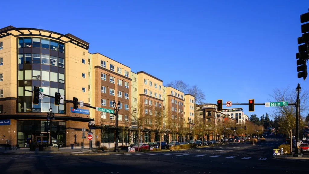 A modern multi-story apartment building with ground-floor businesses is located at a street intersection with traffic lights and a Main St sign under a clear blue sky.