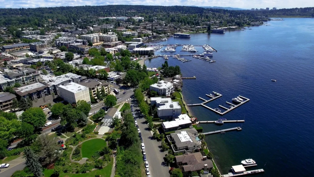 Aerial view of a waterfront city with residential buildings, a marina with boats, green spaces, and a tree-lined shoreline.