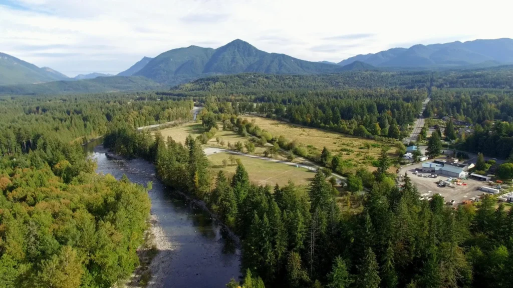 A river runs alongside a forested area and open fields, with mountains in the background and buildings visible on the right side.