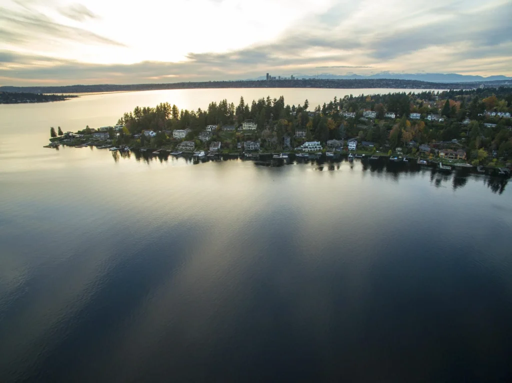 Aerial view of a calm lake with houses along a tree-lined shoreline under an overcast sky at sunset.