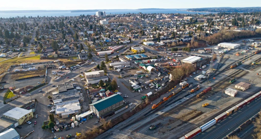 Aerial view of a suburban area with houses, industrial buildings, and a rail yard with multiple freight trains and tracks. Trees and water are visible in the background.