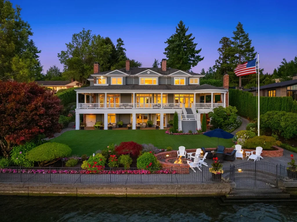 Large waterfront house with multiple chimneys and lit windows, landscaped yard, outdoor seating area, dock, and American flag, photographed at dusk.