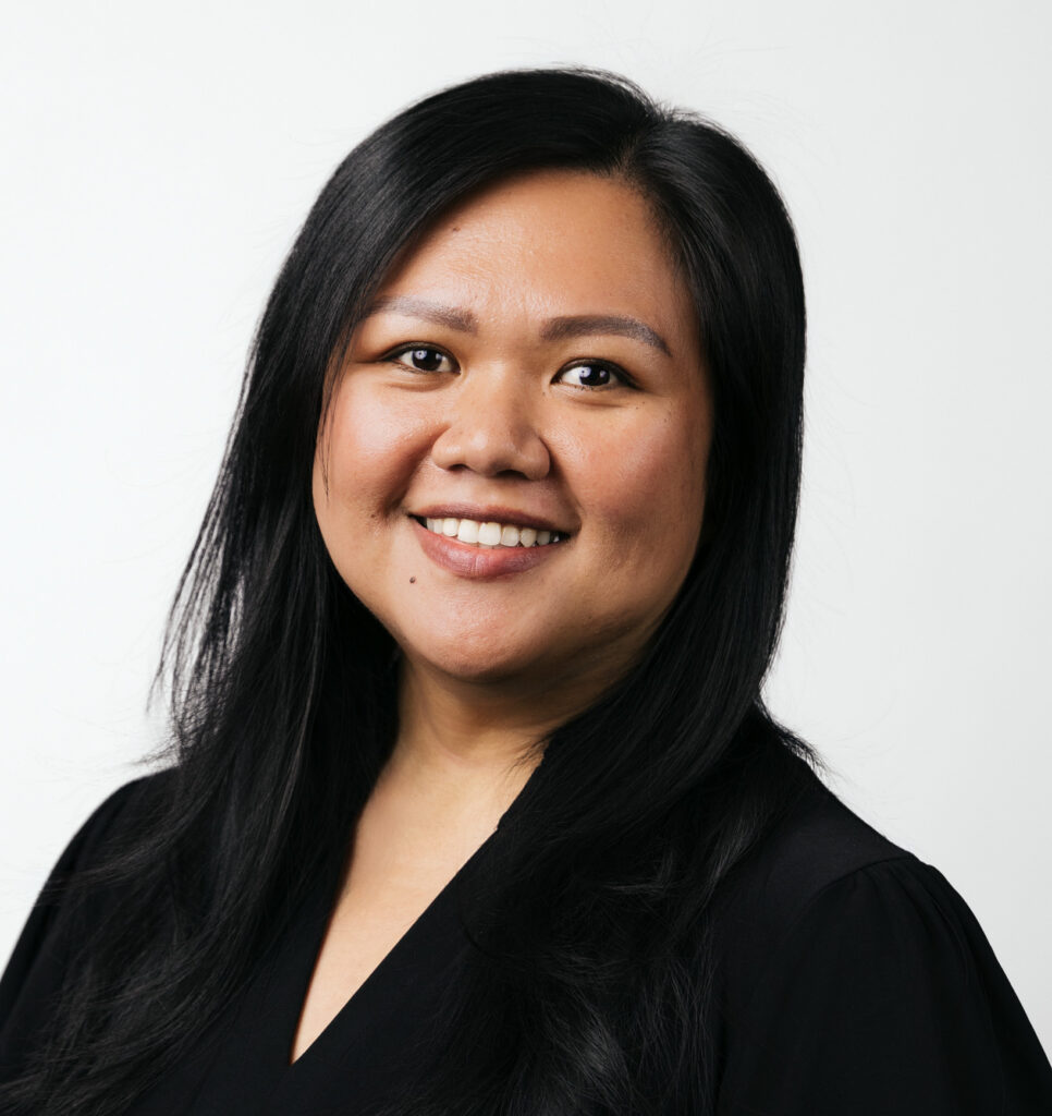 A woman with long black hair wearing a black top smiles at the camera against a plain white background.