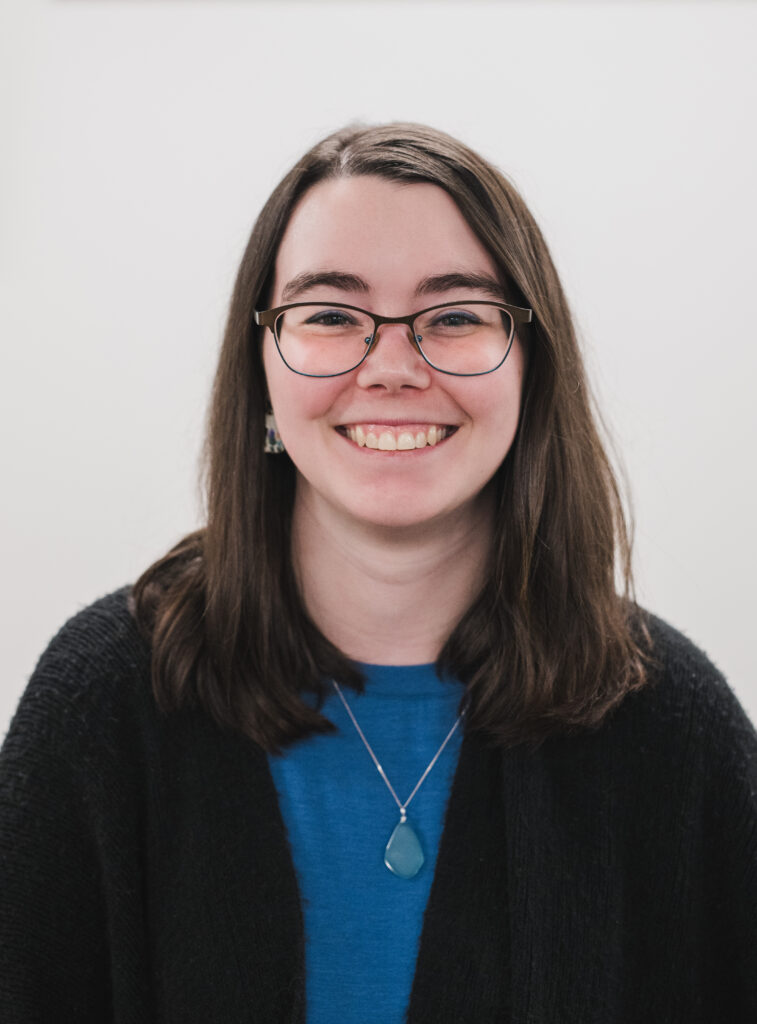 A woman with straight brown hair and glasses smiles at the camera. She is wearing a blue top, a black cardigan, and a pendant necklace. The background is plain and white.