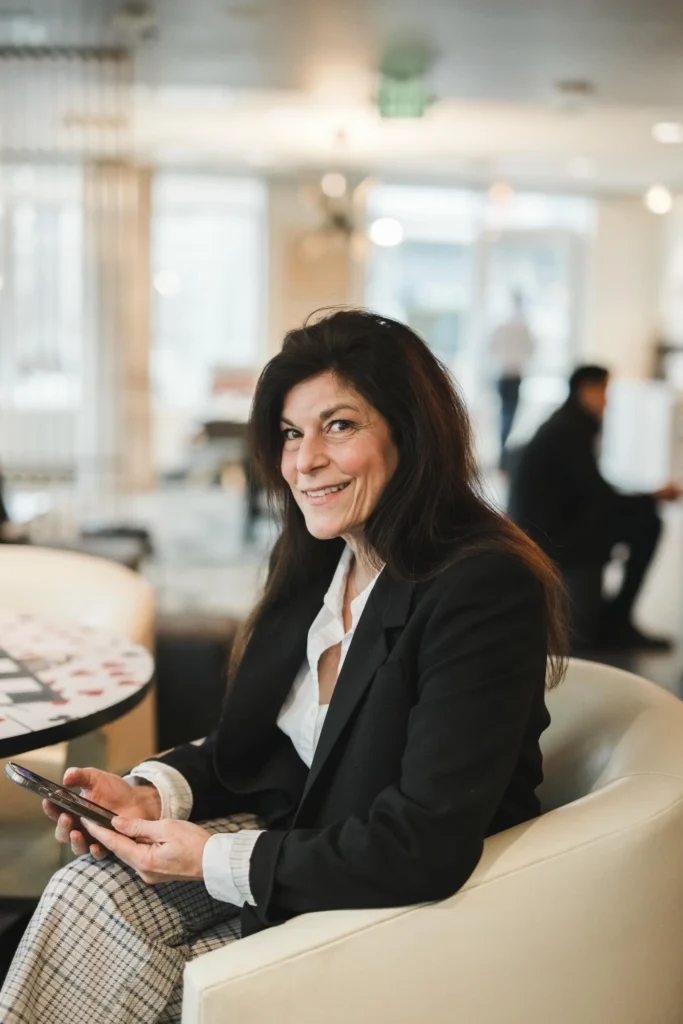 A woman with long dark hair in a black blazer sits in a modern office lounge, holding a smartphone and smiling at the camera.