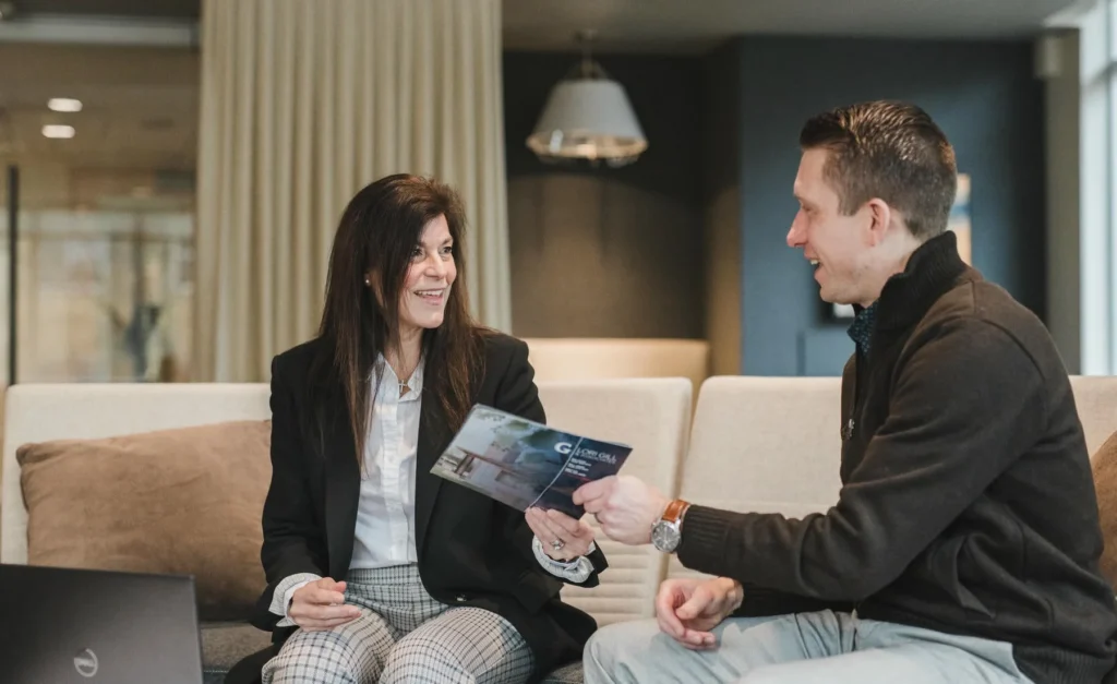 Two people sit on a couch having a conversation; the woman holds a brochure and smiles as she shows it to the man, who appears engaged and attentive.