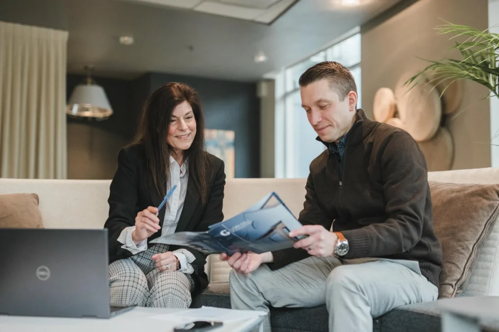 Two people sit on a couch in an office, reviewing documents together. A laptop is on the table in front of them.