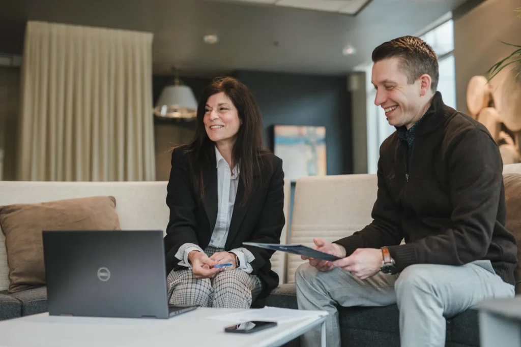 Two people sit on a couch in an office setting, smiling and talking, with a laptop open on the table in front of them.