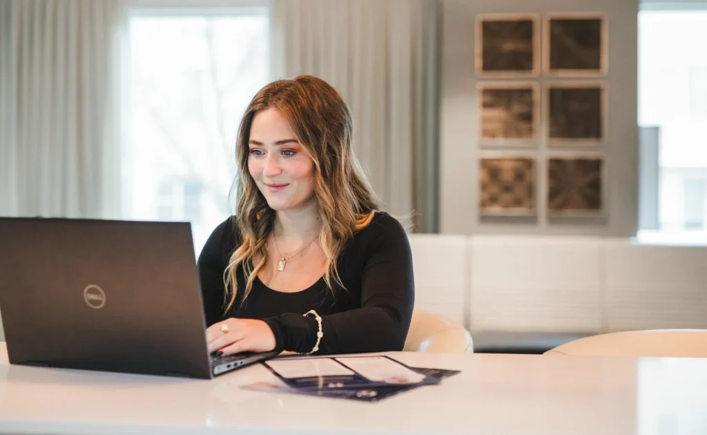 A woman sits at a white table, working on a Dell laptop with documents in front of her, in a bright, modern room.