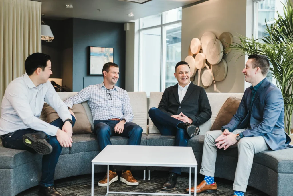 Four men in business attire sit on a curved gray sofa in a modern office, engaged in conversation around a white coffee table.